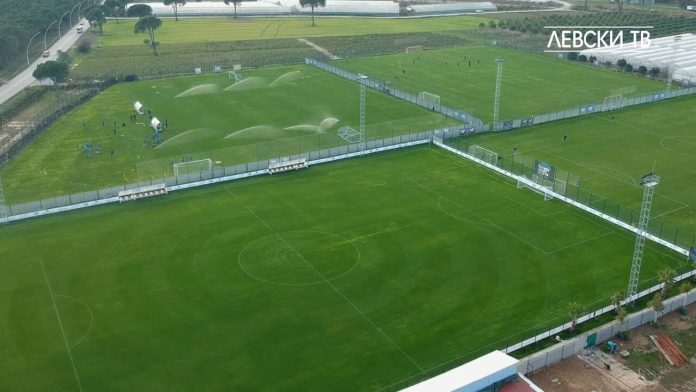 Aerial view of a soccer field being watered by multiple sprinklers, with players and staff on the nearby practice area and dugouts along the sideline.