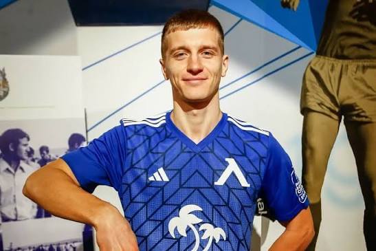 Male athlete in a blue geometric-patterned jersey with a white palm tree emblem, standing with hands on hips and smiling.