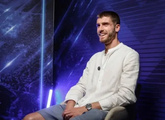 Петко Христов очаква победа срещу ЦСКА Man in a white button-down shirt sits on a chair on a blue neon-lit studio stage, smiling confidently.