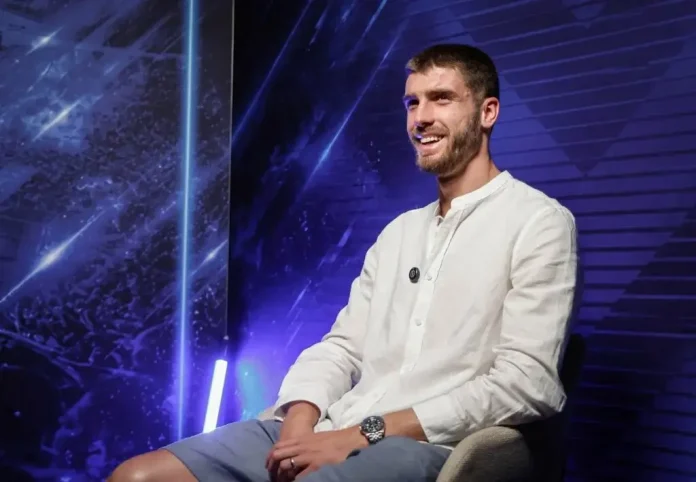 Man in a white button-down shirt sits on a chair on a blue neon-lit studio stage, smiling confidently.