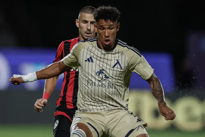 Soccer players contesting the ball during a night match; foreground player in a beige striped kit (Palms Bet sponsor) stretches out as another player in red and black closes in from behind.