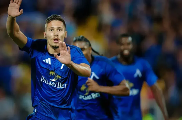 Soccer player in a blue kit with Palms Bet logo raises hands in celebration on the field, teammates behind him.