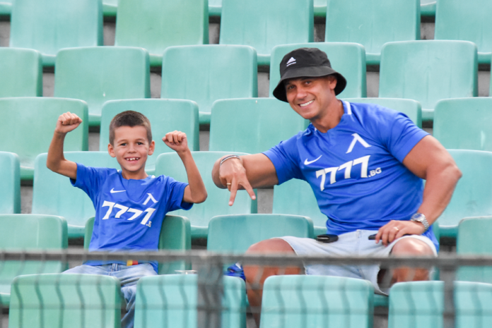 Man and boy in blue shirts flexing their arms while sitting in green stadium seats.