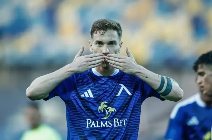 Soccer player in blue jersey kissing his hands on a field, Palms Bet sponsor logo visible on shirt.