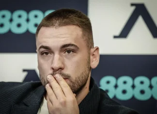 Син шеф с послание към „синята“ общност Man with short hair and stubble rests fingers against his lips at a press event, backdrop shows teal logos and dark panels.