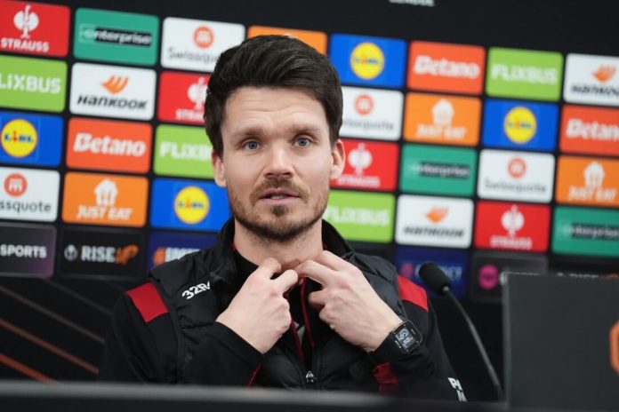 Man at a press conference wearing a black jacket with red accents, hands near his chin, backdrop filled with sponsor logos? and microphones nearby.