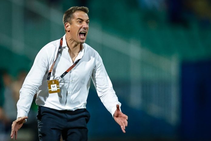Man in a white shirt shouts with arms outstretched on a sports field, wearing a lanyard badge