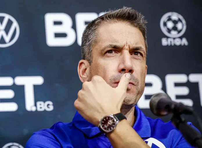 Close-up of a man in a blue polo, finger resting on his lips in a thoughtful gesture during a press conference, microphone nearby.