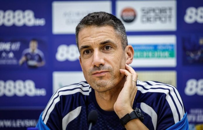 Man in a blue sports top at a press conference, resting his hand on his cheek against a blue sponsor board backdrop.