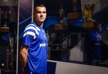 Александър Колев: клубове, участия и голове Male athlete in a blue jersey stands in front of a glass trophy case filled with awards beneath blue lighting.