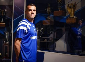 Александър Колев: клубове, участия и голове Male athlete in a blue jersey stands in front of a glass trophy case filled with awards beneath blue lighting.