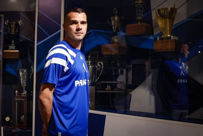 Male athlete in a blue jersey stands in front of a glass trophy case filled with awards beneath blue lighting.