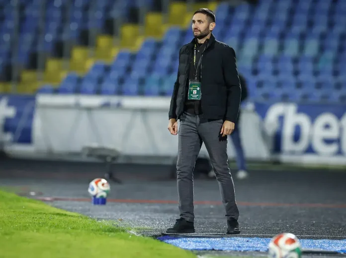 Male coach standing on a sideline of a football pitch, wearing a black jacket and grey pants with a lanyard badge, stadium seats behind him.