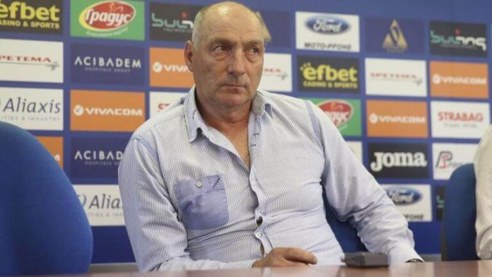 Middle-aged man in a light blue striped shirt sits at a table during a press conference, backdrop covered with sponsor logos.