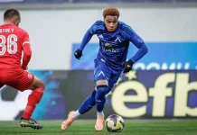Армстронг Око-Флекс: отбори, кариера и награди Soccer player in blue kit dribbles the ball past a defender in red on a green pitch.