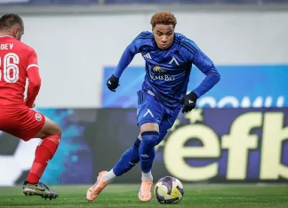Армстронг Око-Флекс: отбори, кариера и награди Soccer player in blue kit dribbles the ball past a defender in red on a green pitch.