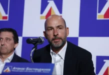 Атанас Бостанджиев се очаква на трибуните за мача на Левски с НЕК Man with a shaved head and gray beard speaks into a microphone at a press conference, with another man beside him and a blue logo backdrop.
