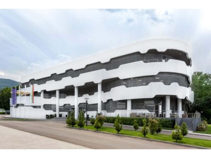 Modern white multi-story building with a curved, layered exterior and columns, surrounded by trees and a paved area.