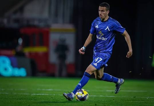 Soccer player in a blue kit dribbling a yellow and purple ball on a grass field during a match.