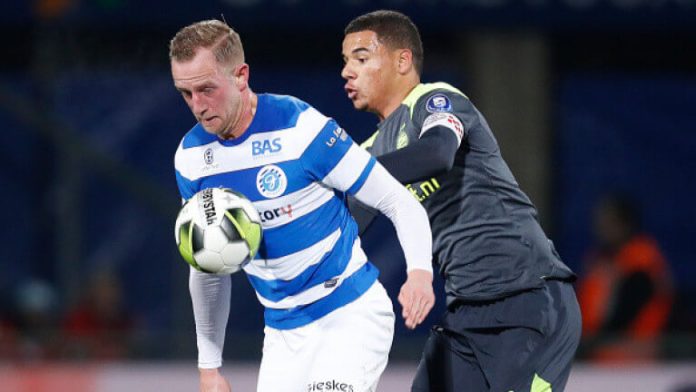 Two soccer players duel for the ball during a match, one in a blue-and-white striped jersey and the other in a dark kit.