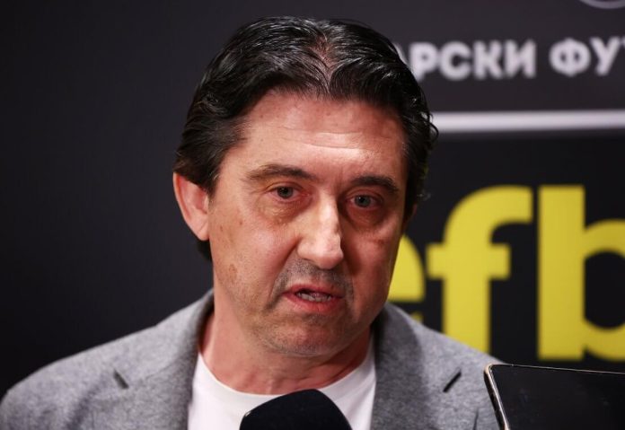 Middle-aged man in a gray blazer and white shirt speaks into a handheld microphone at a press event, with a dark backdrop and partial yellow text in the background.