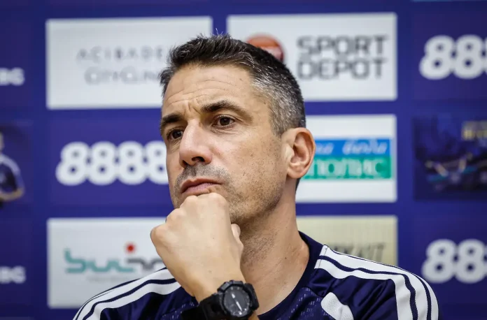 Man in a press conference setting, resting chin on hand, wearing a dark blue athletic jacket and a black wristwatch, serious expression against a blue sponsor wall.