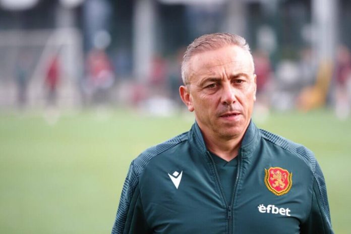 Football coach in a dark green jacket with a crest, standing on a training field.