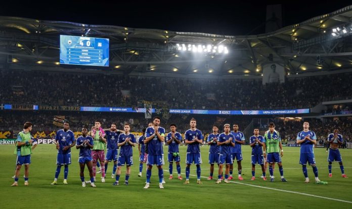 Soccer team in blue uniforms standing in a line on a green field as fans watch from the stands at night.