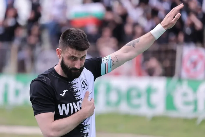 Football player with black-and-white jersey raising his right arm in a stadium, crowd blurred in the background.