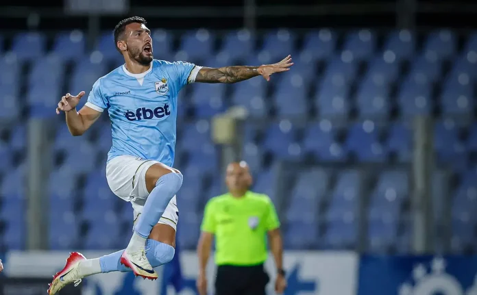 Soccer player in light blue kit jumps mid‑air with arms outstretched during a match on a blue‑seated stadium background.