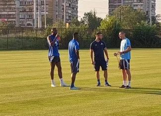 Добри новини за Левски и Хулио Веласкес Coach in a light blue shirt talks to three players in blue uniforms on a sunny outdoor soccer field with buildings in the background.
