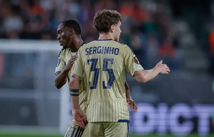 Two soccer players in beige kits embrace on the field after a moment in the match, one showing the back with 'SERGINHO' and the number 17.