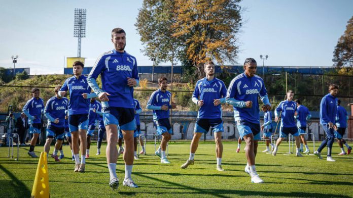 Professional soccer players in blue training outfits warm up on a sunny practice field, moving through cones.