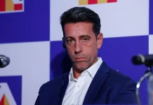 Еду Гаспар води разговори с бразилци Man in a navy suit and white shirt sits at a press event, wearing an earpiece, against a blue checkerboard backdrop.