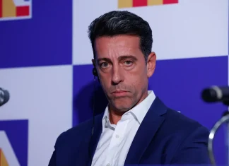 Еду Гаспар води разговори с бразилци Man in a navy suit and white shirt sits at a press event, wearing an earpiece, against a blue checkerboard backdrop.