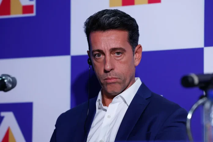 Man in a navy suit and white shirt sits at a press event, wearing an earpiece, against a blue checkerboard backdrop.
