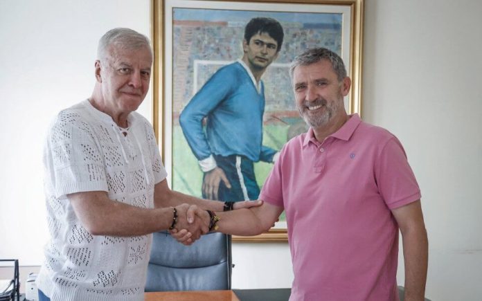 Two men shake hands in an office, standing in front of a framed painting.