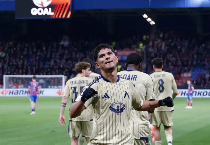 Soccer player in beige striped kit celebrates with fists raised on the field as teammates stand behind him and fans cheer in the stands.