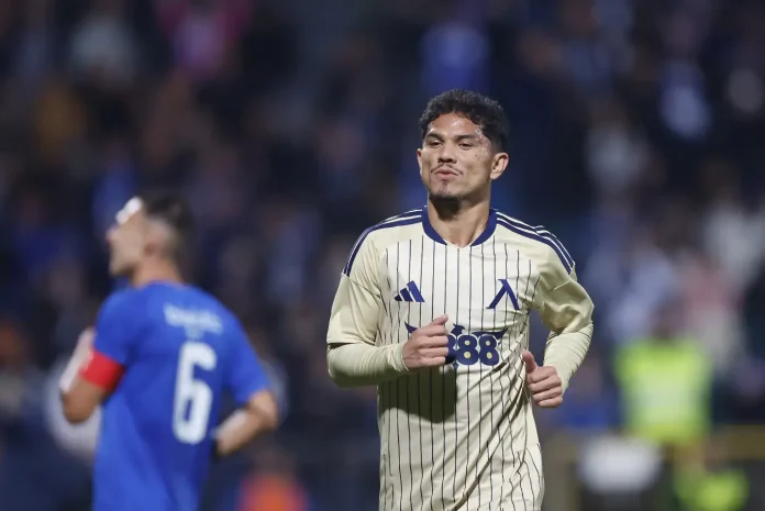Soccer player in a cream striped jersey running on the field during a match, with a blurred blue-clad opponent in the foreground