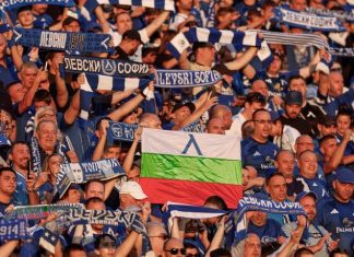 Фенове на Левски с призив преди НЕК: Без излишно напрежение и драми Crowd of football fans in blue and white, holding scarves; a flag with white-green-red and a blue emblem visible in the center