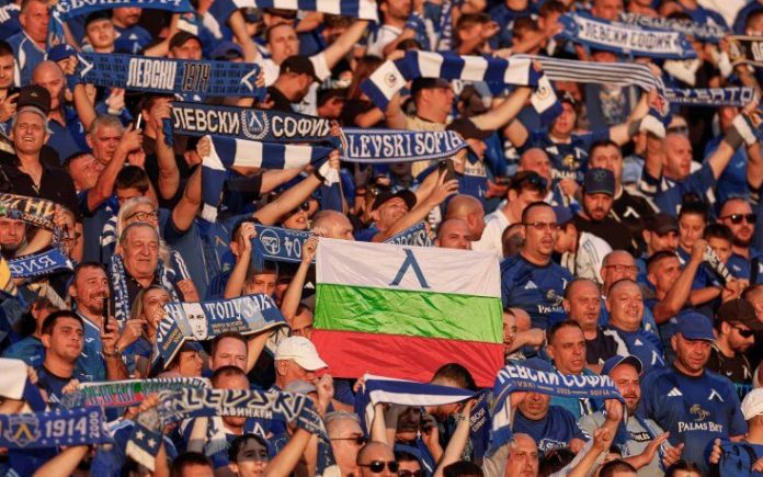 Crowd of football fans in blue and white, holding scarves; a flag with white-green-red and a blue emblem visible in the center