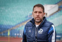 Георги Чиликов преди сблъсъка с Левски: Дунав няма да се предаде без битка Portrait of a man with short brown hair and a beard wearing a navy quilted jacket with a white emblem, standing on a track in a stadium with green seats in the background.