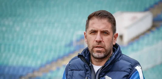Георги Чиликов преди сблъсъка с Левски: Дунав няма да се предаде без битка Portrait of a man with short brown hair and a beard wearing a navy quilted jacket with a white emblem, standing on a track in a stadium with green seats in the background.