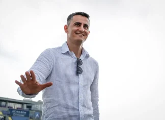 Георги Костадинов каза кога се завръща Бурас Smiling man in a light blue striped shirt extends his hand toward the camera, sunglasses hanging from his chest, outdoors.