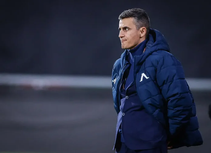 Side profile of a male coach in a blue puffer jacket standing on a field, looking to the right.