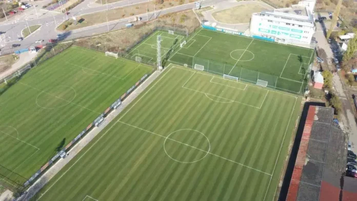 Aerial view of three soccer fields with well-marked lines and a white multi-story stadium at the top-right corner.