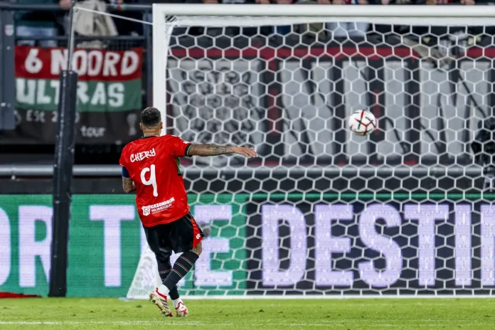 Soccer player in a red jersey number 9 takes a shot toward the goal as the ball is in the air near the net.