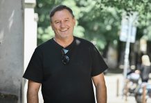 Гошо Гинчев похвали Левски и Веласкес: „Сините“ вече играят като европейски отбор Smiling man in a black t-shirt stands outdoors near a column on a sunny sidewalk, sunglasses hanging from his shirt.