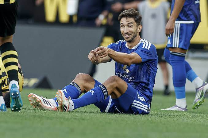 Soccer player in a blue kit sitting on the grass and smiling, gesturing with his arms during a match, with a opponent in yellow and black nearby.