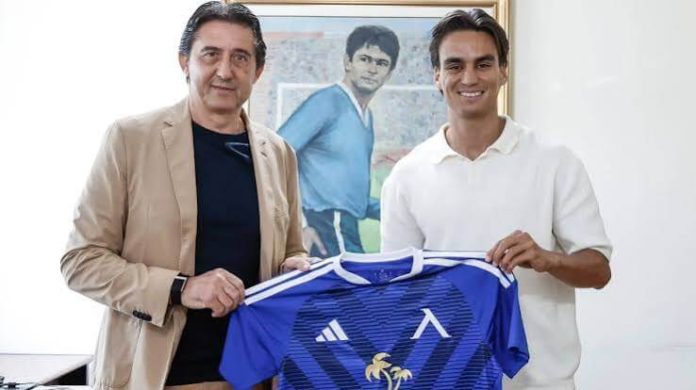 Two men pose indoors, holding up a blue soccer jersey with white accents in front of a painting.